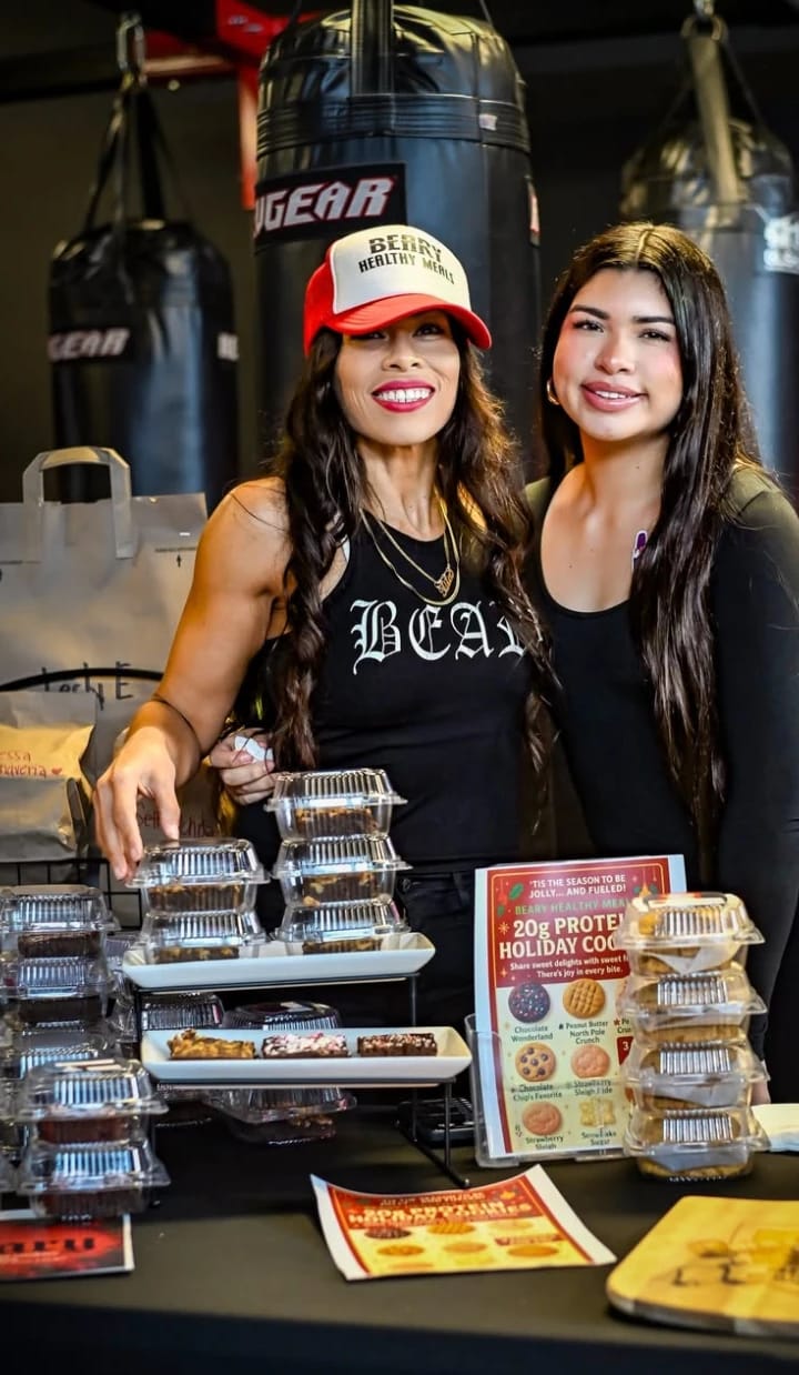 Beatriz in a Beary Healthy Meals hat at her pop-up table in a boxing gym, packaged protein treats in front
